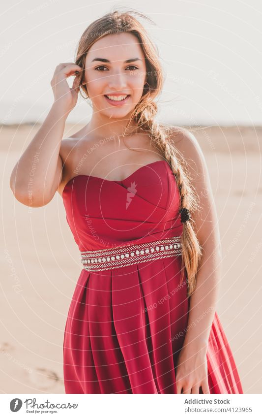 Positive young lady in stylish red dress standing on sandy shore woman coast style charming sensual blond dream romantic freedom positive smile summer
