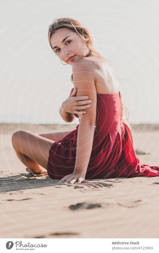 Attractive young lady with long blond hair wearing stylish red dress sitting on coast while looking at camera woman beach sensual sand style summer alone