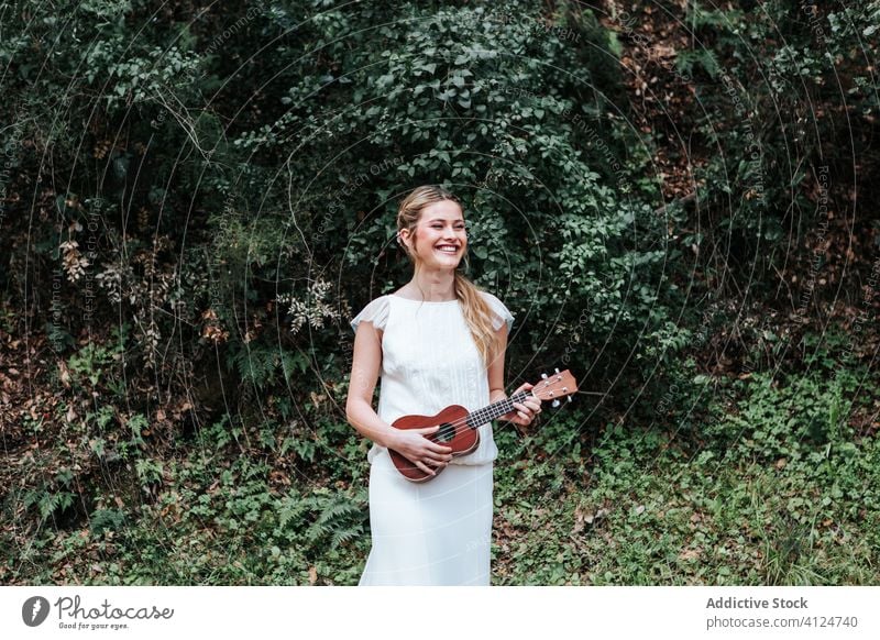 Cheerful bride playing ukulele near bushes woman countryside smile wedding young music female happy dress green cheerful joy summer nature glad tune melody lady