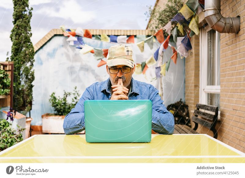 Pensive senior man in glasses having hot drink and using laptop on terrace of country house pensive read watch mug thoughtful device coffee internet male