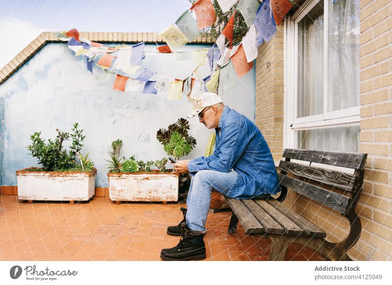 Thoughtful gray haired man in denim clothes using smartphone while chilling alone in garden surfing terrace message read relax pensive senior use rest browsing