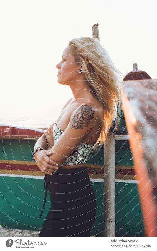 Tattooed female standing near boat on sandy beach with arms crossed woman style tropical sunset tattoo slim hand raised head shore water coast blond wave admire