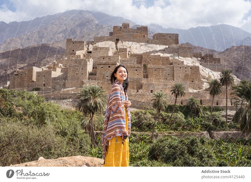 Cheerful ethnic woman standing against ancient village and mountains happy exotic traveler historic stone tropical ornament heritage nature old green palm