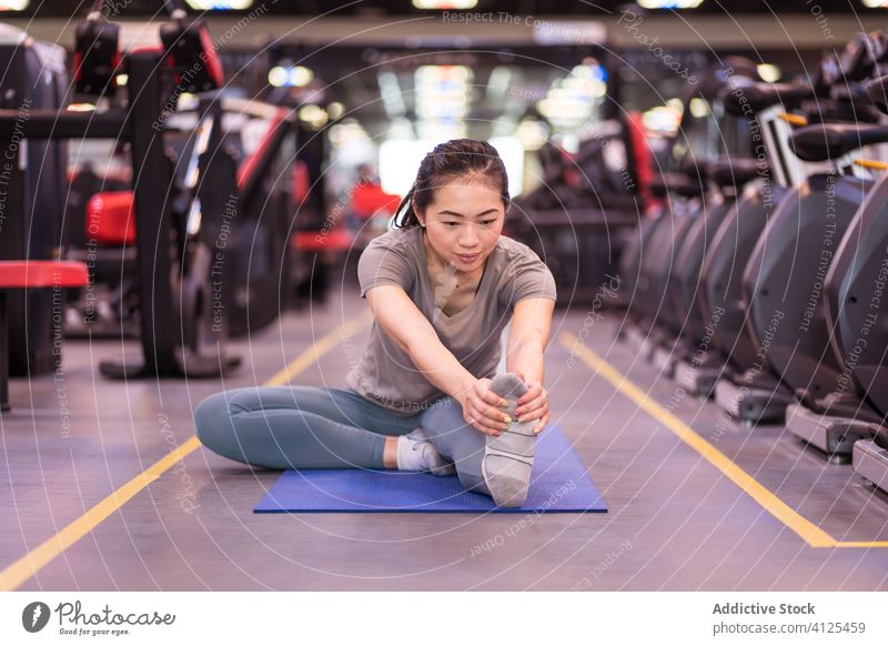 Strong young ethnic sportswoman stretching leg on mat in gym training fitness warm up club slim slender wellness exercise workout athlete sportswear asian