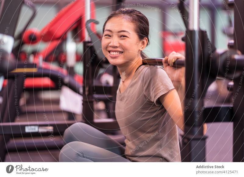 Positive Asian sportswoman leaning on barbell in modern gym and looking at camera rest workout machine content relax proud confident ethnic asian break fitness