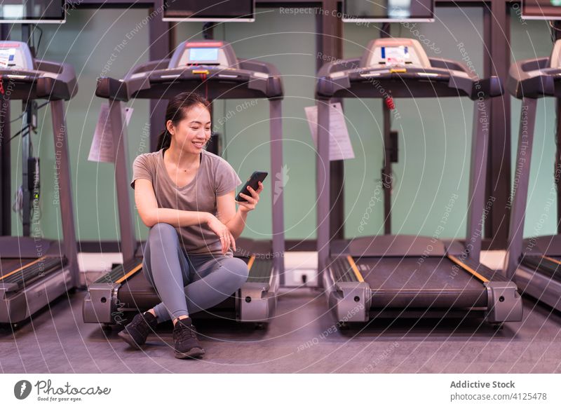 Cheerful young ethnic sportswoman sitting on treadmill and using mobile phone in gym smartphone rest break workout cardio recovery cheerful social media