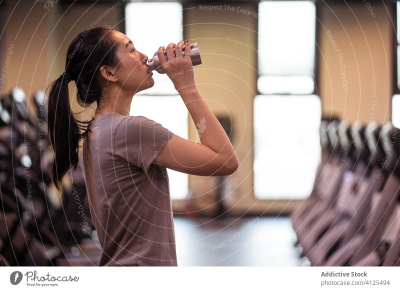 Young ethnic sportswoman drinking water from bottle in gym fresh workout thirst hydrate athlete fitness break young slim sportswear rest training relax activity