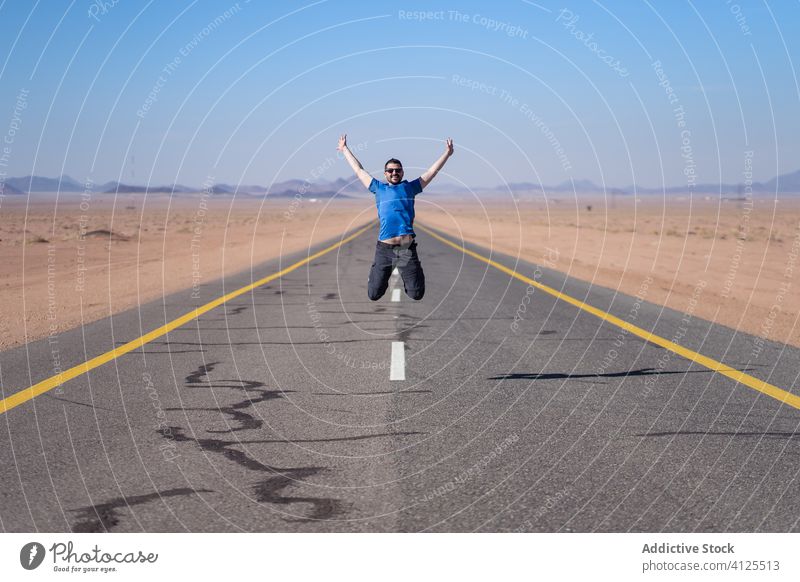 Excited young man jumping on asphalt road excited adventure freedom travel desert leap saudi arabia male happy cheerful arms raised celebrate casual cloth