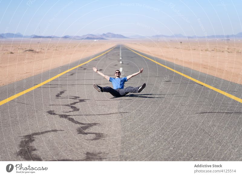 Cheerful man sitting on road mountain freedom endless route desert gesture countryside road marking positive content peace jeans daytime asphalt optimist