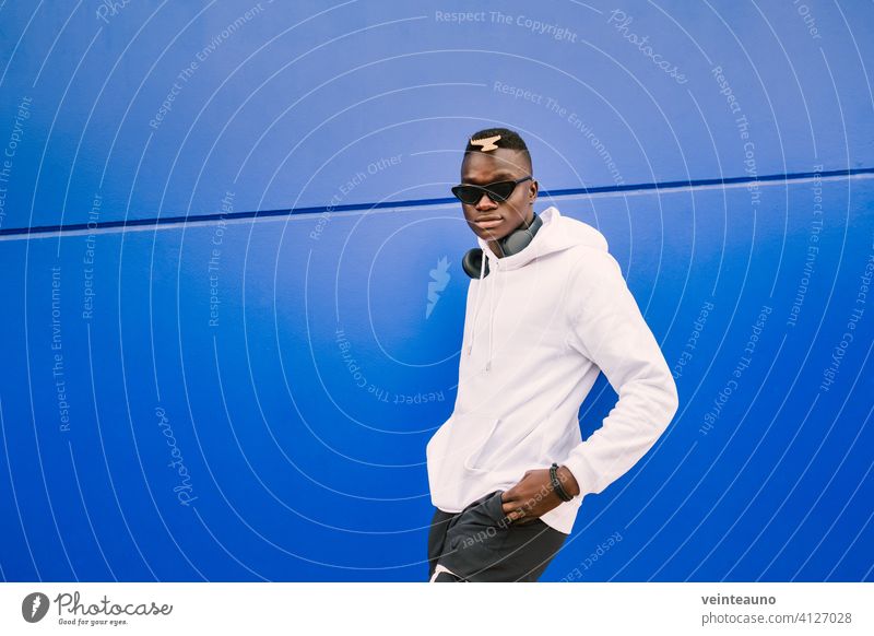 Young african black man against a blue wall looking at camera wearing a white sweatshirt and sunglasses with style and confidence young afro street background
