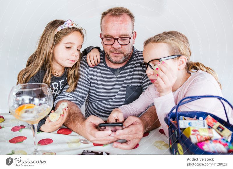 Father and daughters using smartphone at table father banana children sibling eat together home man girl little browsing device kid gadget snack social media