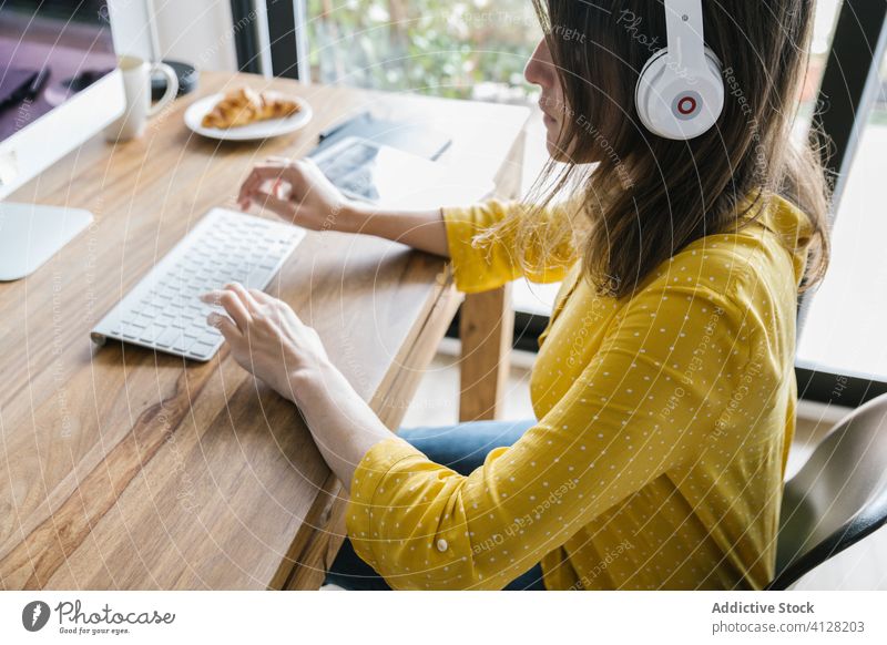 Concentrated woman using computer at home keyboard telework desk pc headphones freelance typing tablet croissant coffee browsing online gadget cup workplace
