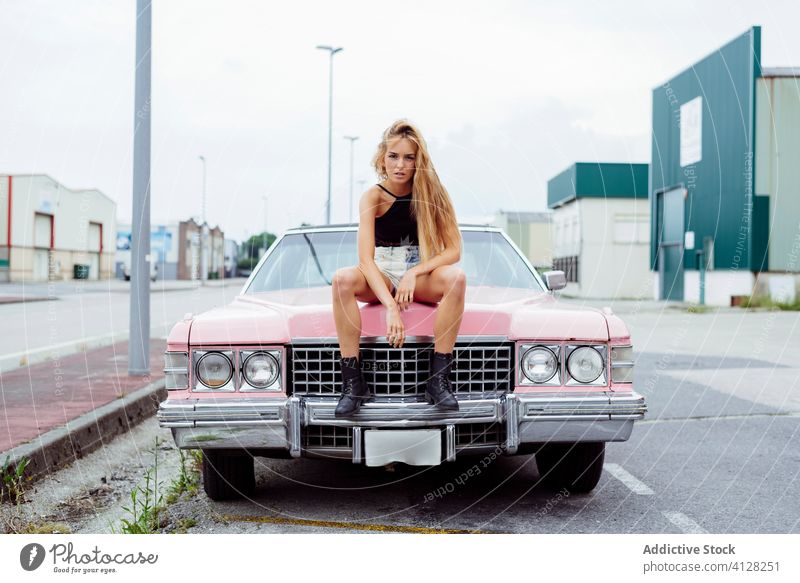 Attractive blonde girl sitting on the hood of a classic pink car on the street woman young braids old grunge summer portrait leisure urban city outskirts