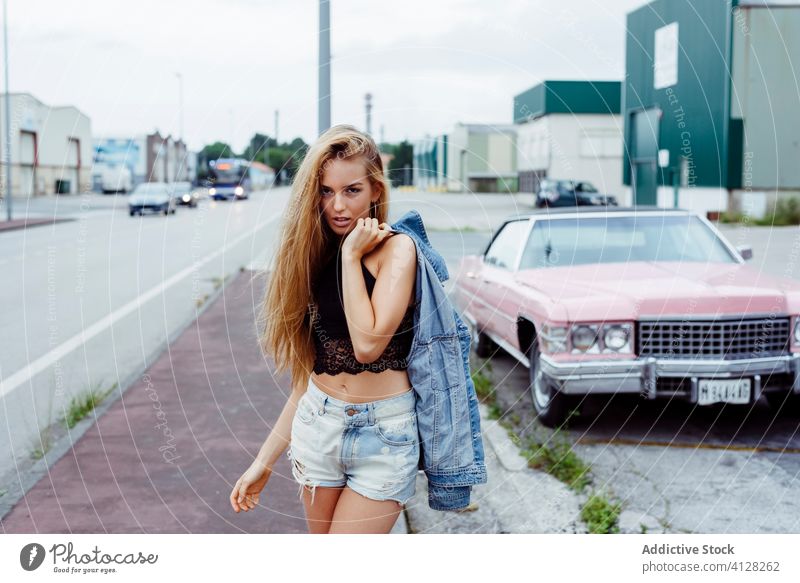 Sensual blonde girl standing on the sidewalk looking at the camera near to a classic pink car woman young braids sitting old grunge summer portrait leisure