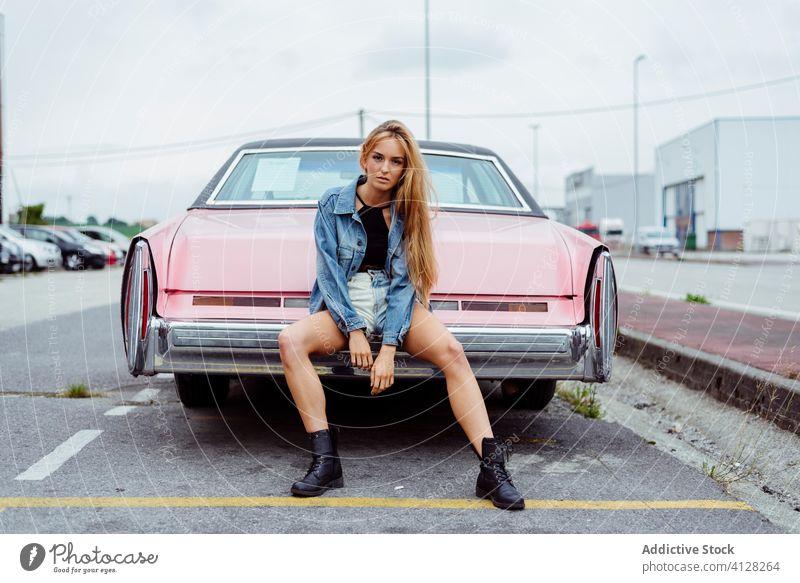 Sensual blonde girl sitting on the bumper of a classic pink car woman young braids sidewalk old grunge summer portrait leisure urban city outskirts attractive
