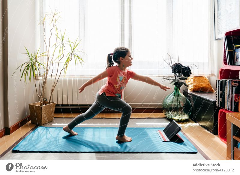 Little girl practicing yoga in warrior two pose kid asana practice home tablet virabhadrasana online flexible stretch little mat balance concentrate video