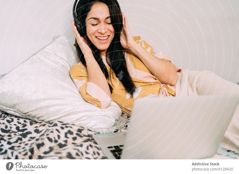 Happy woman with laptop and headphones on bed music listen chill happy enjoy comfort lounge bedroom relax lifestyle rest gadget device home young cozy apartment