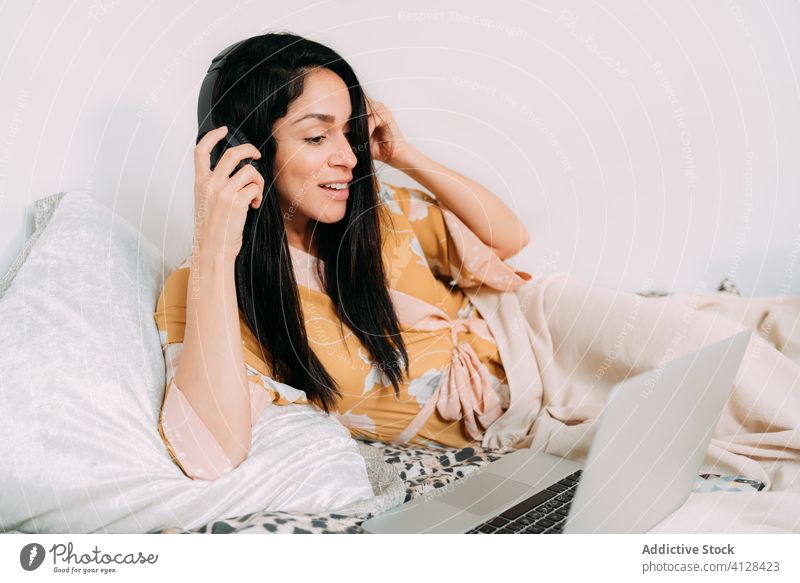 Happy woman with laptop and headphones on bed music listen chill happy enjoy comfort lounge bedroom relax lifestyle rest gadget device home young cozy apartment