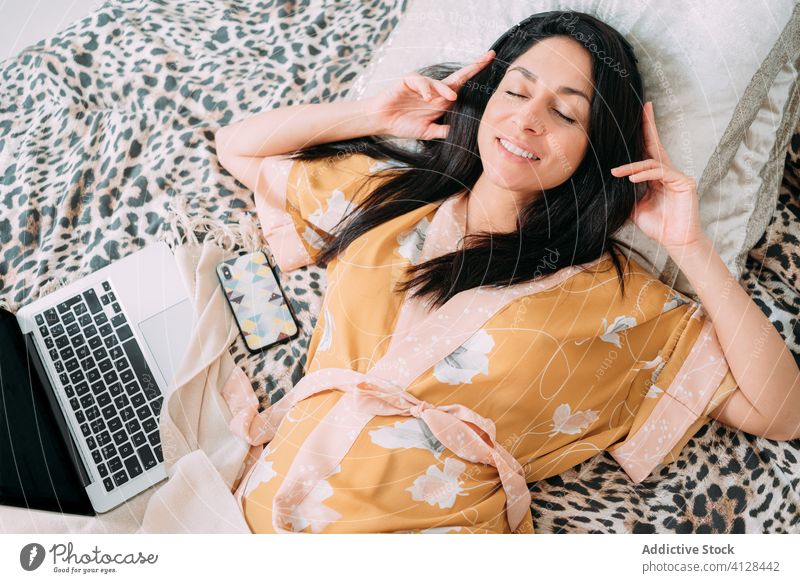 Happy woman with laptop and headphones on bed music listen chill happy enjoy comfort lounge bedroom relax lifestyle rest gadget smartphone device home young