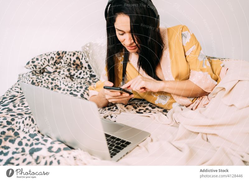 Woman browsing her smartphone while listening to music in bed woman silk robe chill headphones social media comfort communicate using relax internet laptop rest