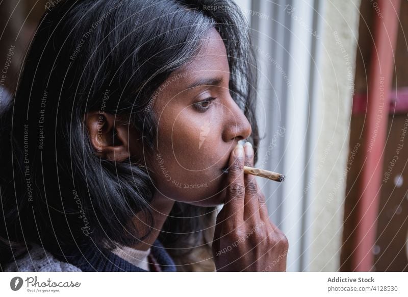 Ethnic woman smoking weed on terrace smoke marijuana relax enjoy roll up young cigarette female ethnic indian hindu casual outfit apparel looking away dope