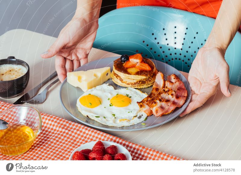 Anonymous crop hands putting plates with delicious fried eggs with bacon and cheese on table for breakfast person cupcakes strawberries black berries honey food