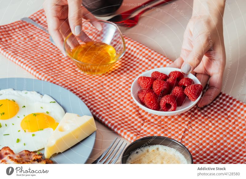 Anonymous crop hands putting plates with raspberry and honey on table for breakfast tasty food morning sweet fresh healthy fruit meal dish yummy snack portion