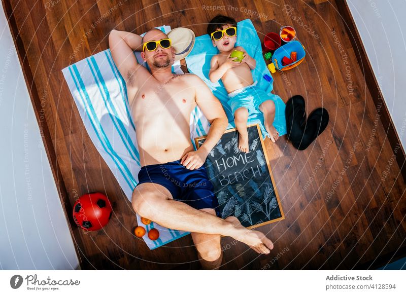 Father and son in sunglasses resting on floor in swimming trunks father beach home self isolation quarantine sunbath dream anticipate social distancing positive