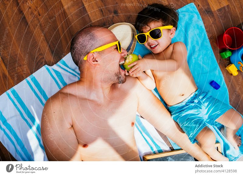 Cheerful kid and father having home beach during quarantine boy cheerful fun social distancing self isolation anticipate sunglasses blue swimwear apple lying