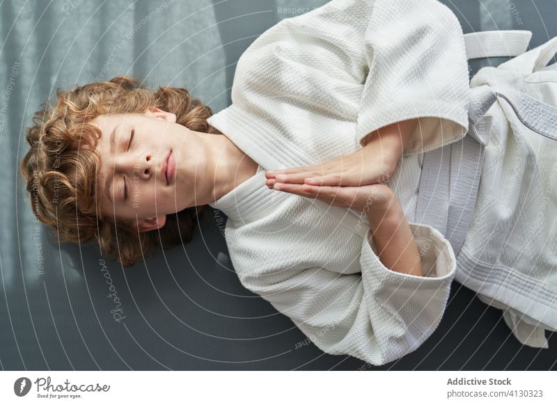 Focused young boy in white kimono practicing meditation practice lying down meditate judo child home eyes closed hands together exercise calm relax wellness