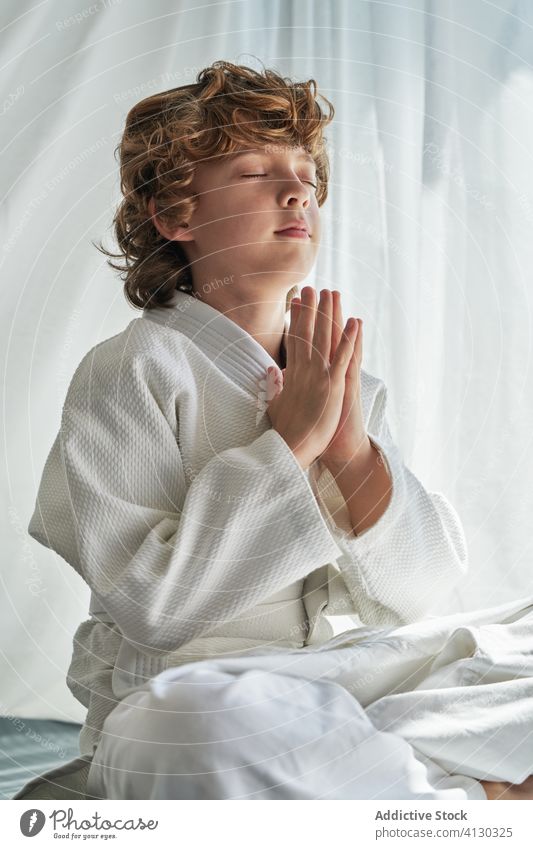 Concentrated young boy in white kimono practicing meditation practice meditate judo child home eyes closed legs crossed hands together exercise calm relax
