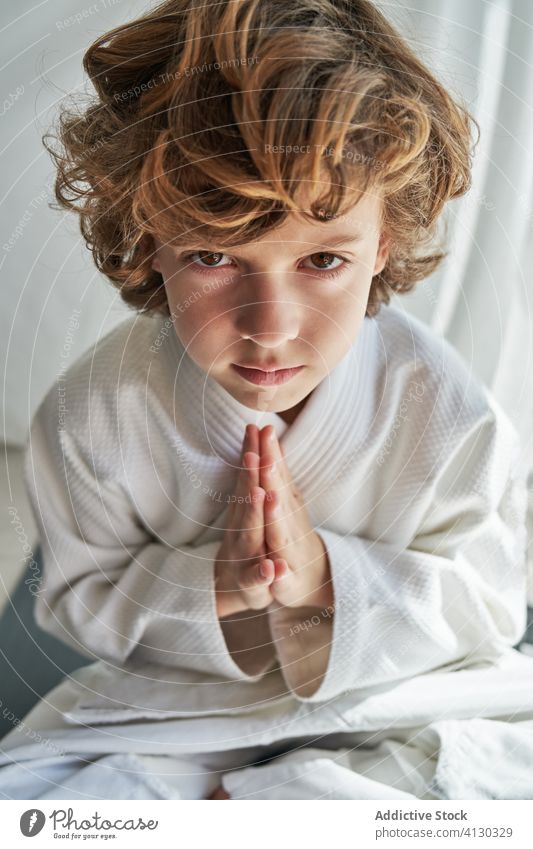 Concentrated young boy in white kimono practicing meditation practice meditate judo child home looking at camera legs crossed hands together exercise calm relax