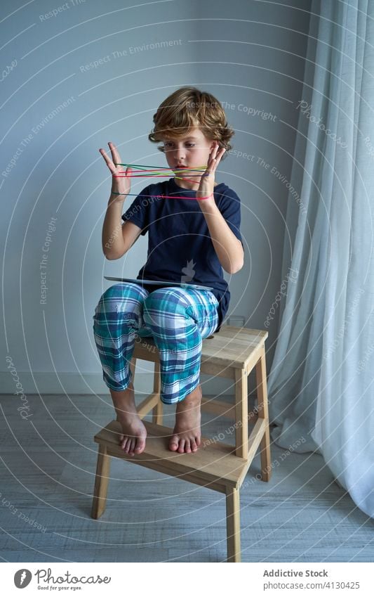 Child with tablet learning cats cradle game kid string play creativity figure stool pajamas home boy focus preteen tie imagination skill develop finger device