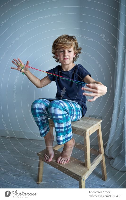 Boy playing brainteaser game with string kid tablet learn cradle creativity figure stool cat pajamas home boy focus preteen imagination skill grimace smile