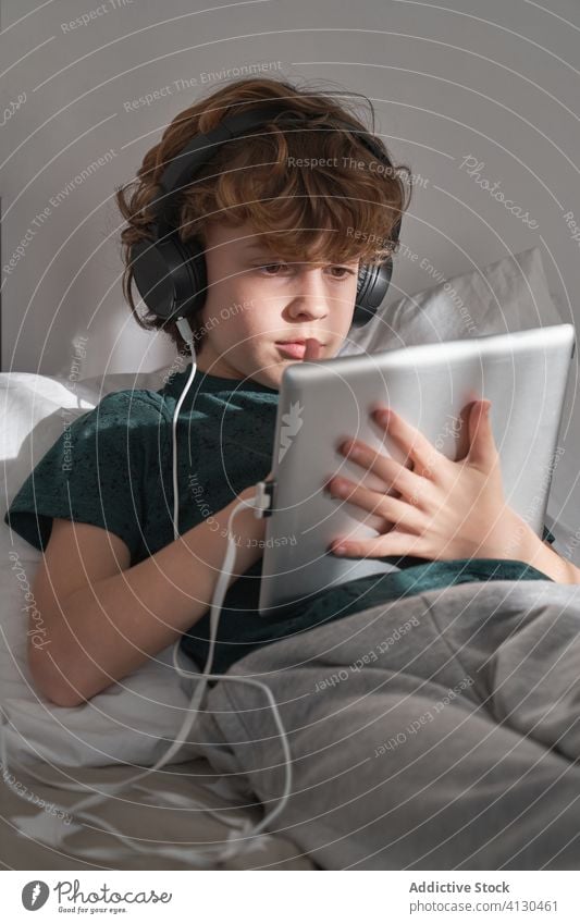 Satisfied child in headphones using digital tablet while chilling in modern bedroom at home in day off use boy kid happy listen relax social media play read