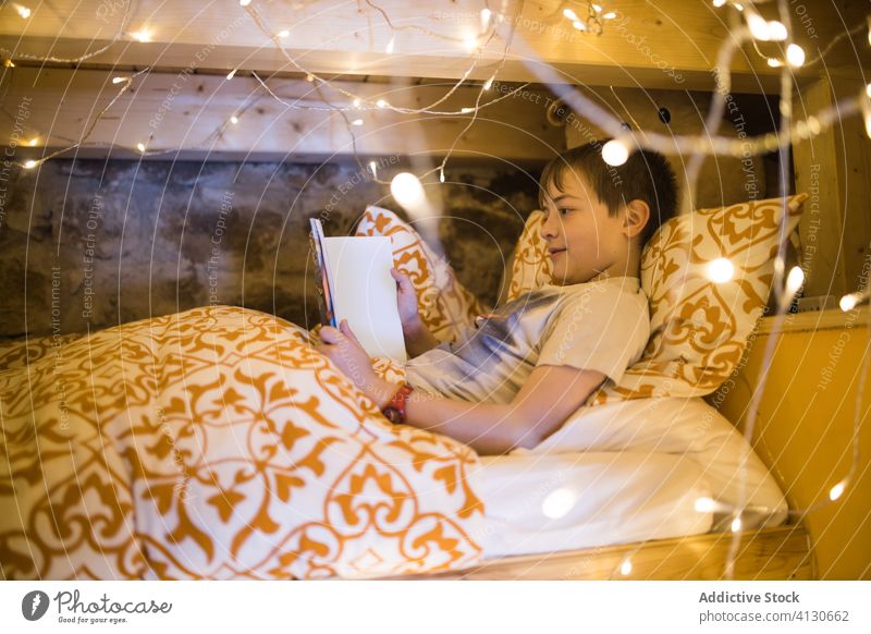 Kid watching tablet in bed decorated with garland boy using cozy comfort video chill leisure home evening glow illuminate bedroom relax rest browsing spain