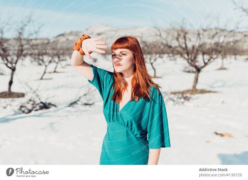 Young beautiful female with long red hair in turquoise outfit looking away while standing in snowy field in sunny day woman style trendy sensual redhead young