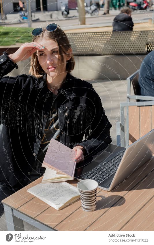 Female student with laptop sitting in street cafe in sunlight freelancer copybook cover eyes bright eyeglasses coffee break free time internet using device