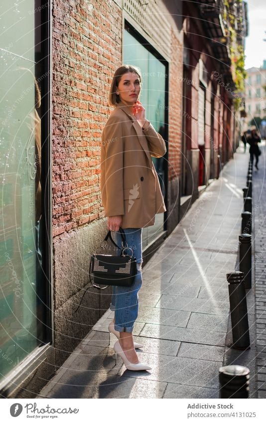Stylish model with handbag standing on pavement in daylight style fashion cloth elegant sidewalk touch chin sunbeam slim thoughtful dreamy charming slender