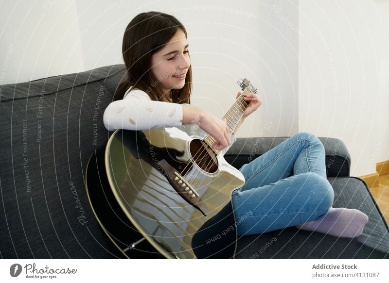 Young smiling girl playing acoustic guitar on sofa at home young cheerful legs crossed relax instrument joy comfort fun hobby music smile leisure cozy melody
