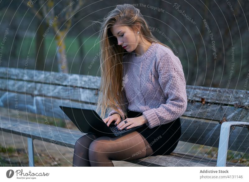 Side view of casual female using laptop on wooden bench in park woman typing email internet device gadget online relax connection rest surfing flowing hair