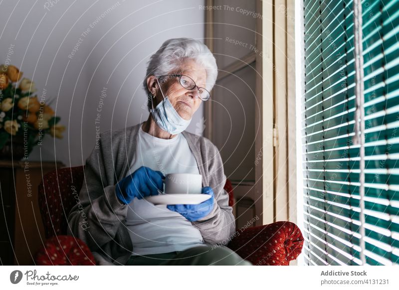 Aged woman with cup of tea staying at home during quarantine coronavirus senior drink window lonely sad pensive mask glove abandoned old female protect alone