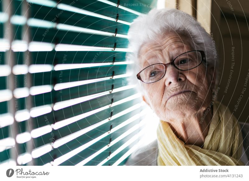 Elderly woman standing near window during pandemic senior coronavirus stay home lonely unhappy sad shutter pensive quarantine prevent old alone depression