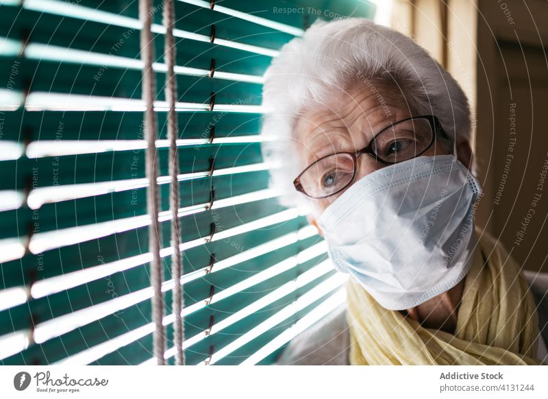 Elderly woman standing near window during pandemic senior coronavirus stay home lonely unhappy sad shutter pensive quarantine mask prevent old female protect