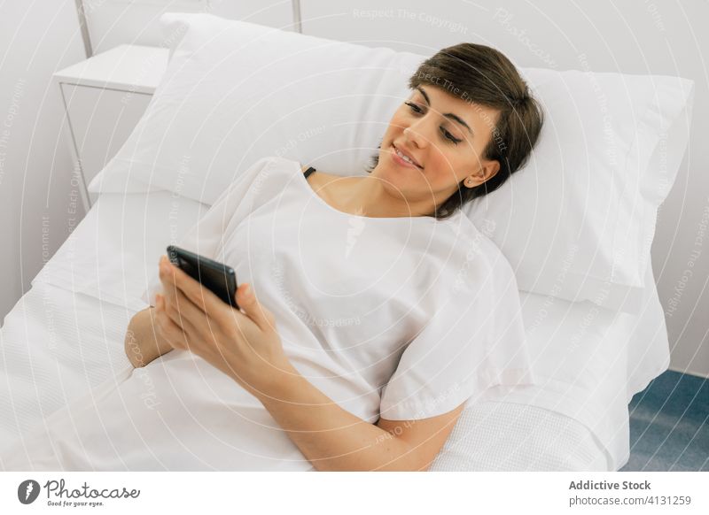 Happy patient using smartphone in hospital ward woman lying down smile chair bed female modern robe cheerful white browsing clinic device texting gadget