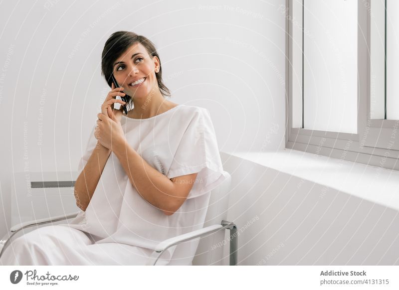 Happy patient talking on a smartphone in hospital ward woman speak using smile chair sit bed female modern robe cheerful white browsing clinic device gadget
