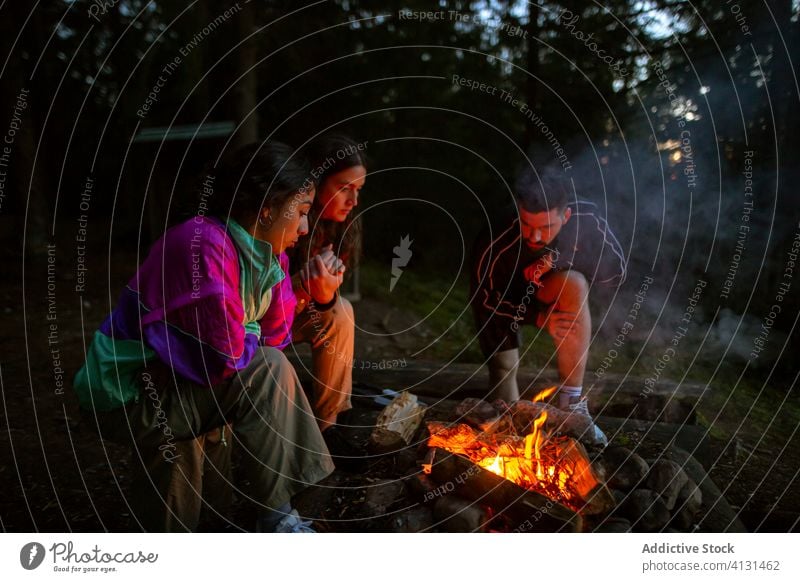 Group of multiethnic friends near campfire in forest group gather bonfire woods warm up evening company diverse multiracial camper relax firewood log campsite