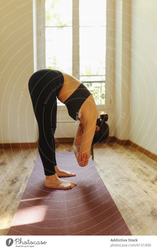 Unrecognizable woman standing in fold pose at home in sunlight yoga apartment calm practice asana mat energy mindfulness barefoot healthy lifestyle activewear
