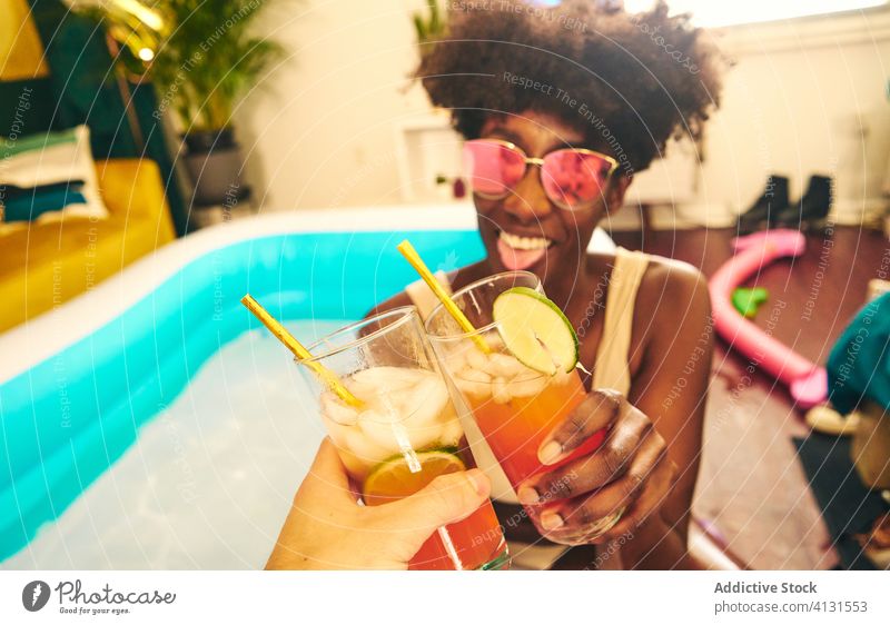 Cheerful ethnic woman clinking glasses of cocktail with friend pool stay at home alcohol drink inflatable self isolation having fun female social distancing