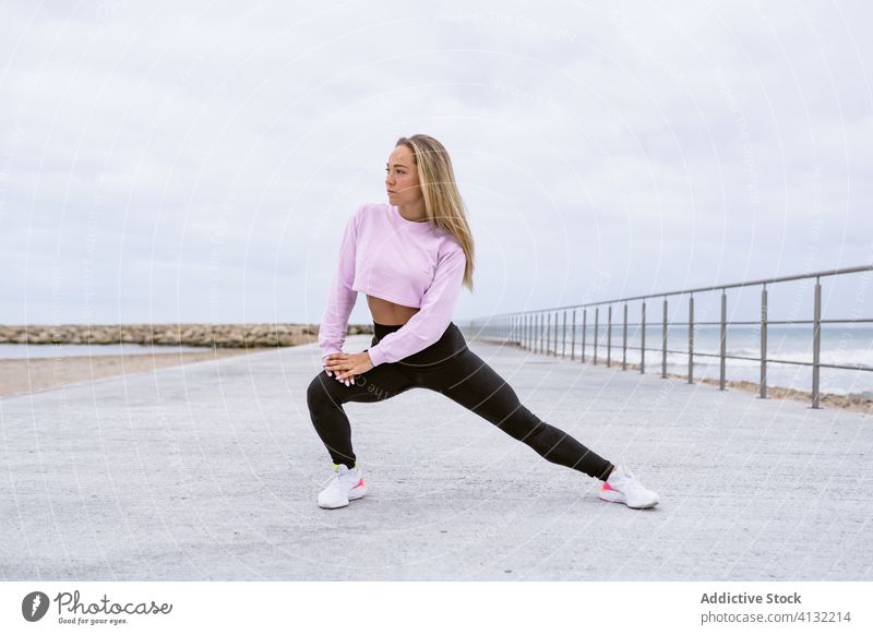 Fit sportswoman doing side lunge on embankment near ocean stretch leg workout activewear fence flexible training warm up sneakers wellbeing sky cloudy activity